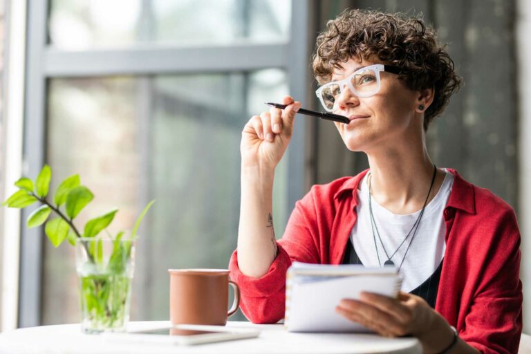 Woman sitting near a window, thinking with a pen near her mouth and notepad in her hand.