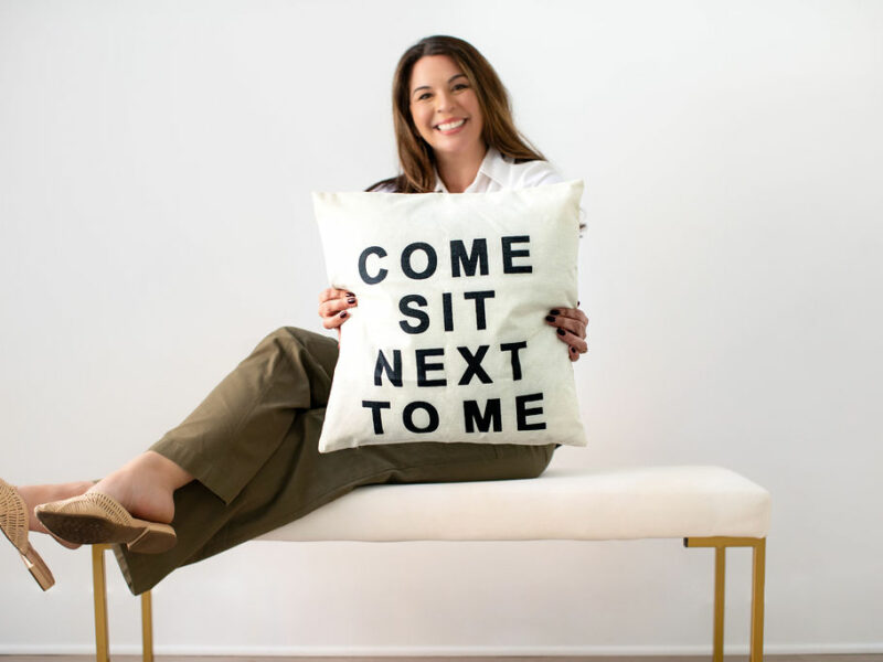AliceAnne Loftus holding a pillow that reads, "Come sit next to me"